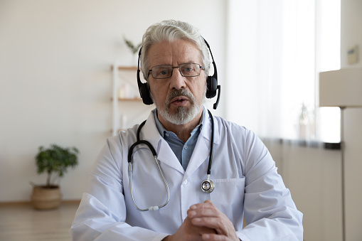 Older male doctor therapist wearing headset videoconferencing talking to web camera consulting virtual patient online by video conference call chat. Telemedicine, telehealth concept. Webcam view.