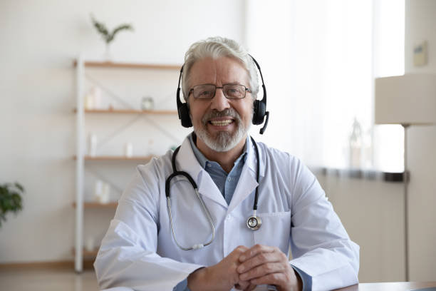 smiling senior doctor wearing headset looking at camera, telemedicine concept - senior adult happiness computer looking at camera imagens e fotografias de stock