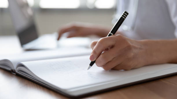 female doctor hand making notes in journal using laptop - médico geral imagens e fotografias de stock