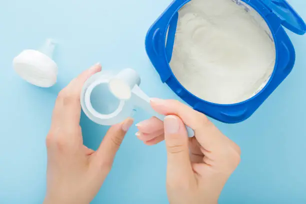 Young mother hands pouring powder of milk formula and mixing with water in plastic bottle. Opened container on light blue table background. Baby feeding. Closeup. Point of view shot. Top down view.