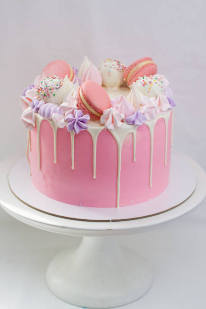 istock Tender pink cake decorated with melted white chocolate, macaroons, meringues, cake pops and candies. Plain background. 1224756369
