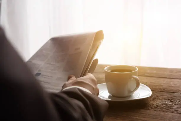 Photo of A man in a business suit in the morning in the sun reads the news in the newspaper, next to a cup of black coffee. Reading business news. Place for text