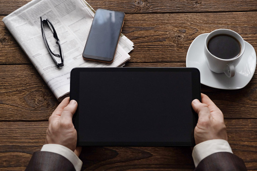 Top view on male hands holding a tablet, a cup of coffee and a morning newspaper. Work is all office. Business concept. Place for text.