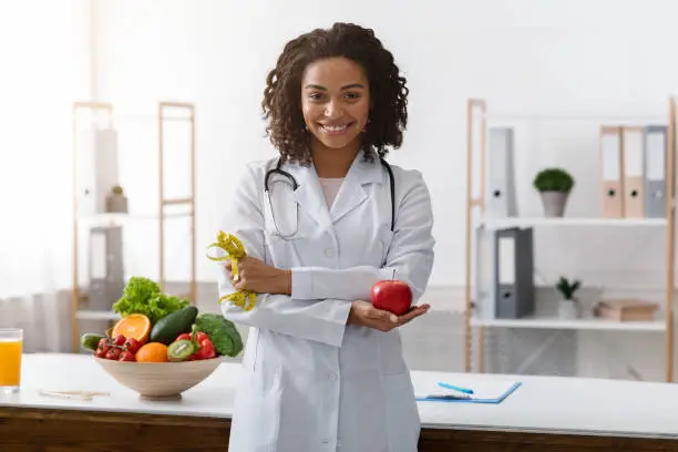 African female dietician with crossed arms holding fresh apple, posing at workplace