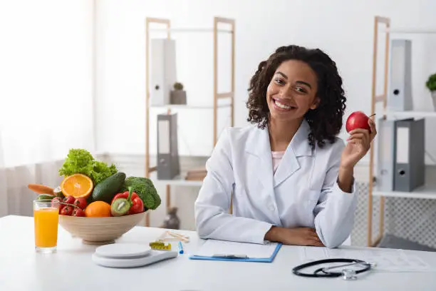 Diet while lockdown. Pretty black lady dietologist holding apple in her hand and smiling, recommending fresh fruits, posing at clinic, copy space