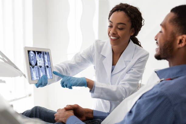 Smiling african female dentist showing patient teeth xray Smiling african female dentist showing young man patient teeth xray, using digital tablet orthodontist stock pictures, royalty-free photos & images