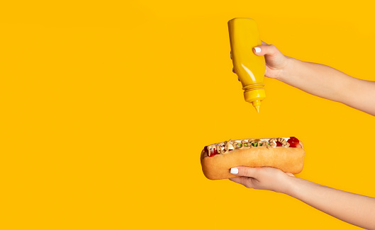 Unrecognizable woman squeezing mustard onto hot dog against orange background, empty space. Panorama