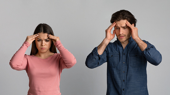 Young couple having headache after quarreling, touching their temples with sad face expressions, standing over light studio background, panorama