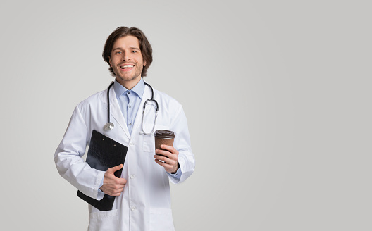 Young smiling doctor holding medical chart and takeout coffee, having break between patients reception, looking at camera over light background