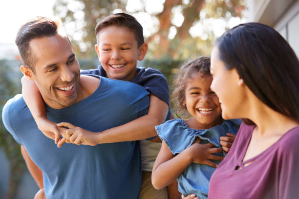 Smiling Hispanic Family With Parents Giving Children Piggyback Rides In Garden At Home Smiling Hispanic Family With Parents Giving Children Piggyback Rides In Garden At Home hispanic stock pictures, royalty-free photos & images