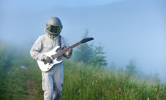 Astronaut in space suit walking on grassy path and playing on guitar. Cosmonaut guitarist with musical instrument strolling down foggy meadow with green grass. Concept of music, astronautics, nature.