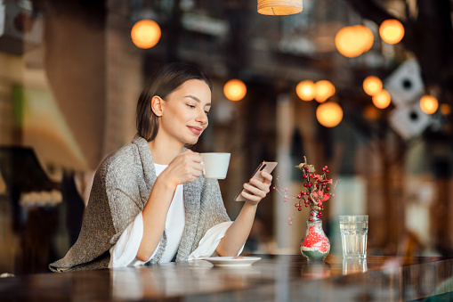 Attractive young woman sitting and relaxing in a cafe, holding a smart phone, sending text messages, smiling and having a cup of coffee