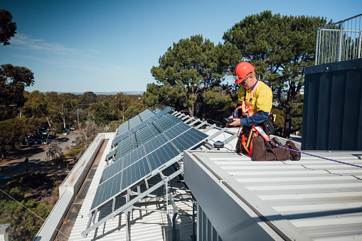 A mature man at work, repairing some solar cell panels on the roof a university. Wearing his overalls, harness and hardhat.