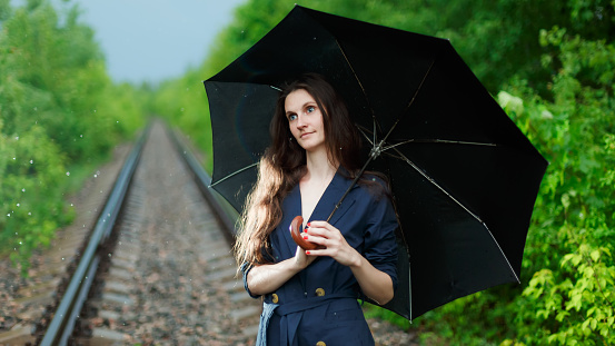 Girl with a black umbrella on the railway tracks in the forest. It's raining. The girl has a blue cloak. The hair is loose. Look away. The girl is lit by bright light. Bright green forest around.
