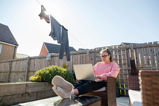A low angle view of a young woman sitting outdoors on a chair in her garden with her feet up while working from home during the COVID-19 pandemic. She is using a laptop.