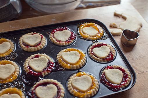 Home baking. Cooked Jam Tarts with heart shaped centres on baking tray. Jam overspilling onto baking tray. Heart shaped cookie cutter with pasty to one side.