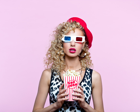 Portrait of surprised blond curly hair woman wearing leopard print top and red beret. Female in 80’s style outfit wearing 3-d glasses, holding popcorn, looking at camera. Studio shot on pink background.