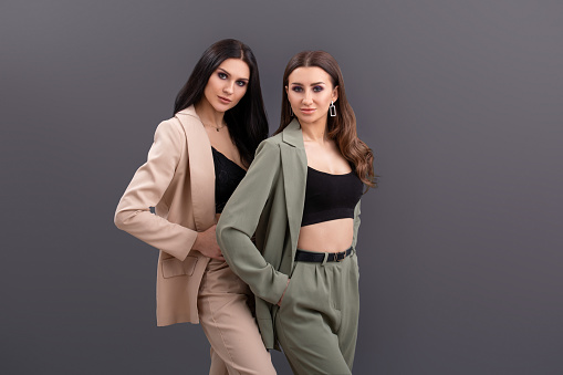 Fashion portrait of Two attractive business women wearing in suits and tops posing on grey background