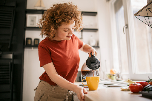 Happy smiling woman with red curly hair pouring herself a cup of coffee early in the morning
