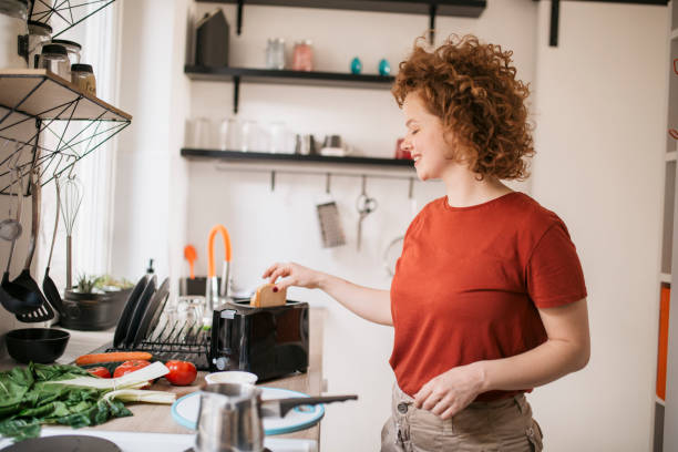 Happy young redhead woman during her early morning ritual Happy smiling woman with red curly hair preparing breakfast for herself, toasting the bread toaster stock pictures, royalty-free photos & images