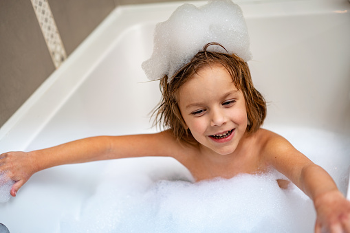 happy infant baby take a bath and playing rubber duck with foam bubbles in bathtub at home