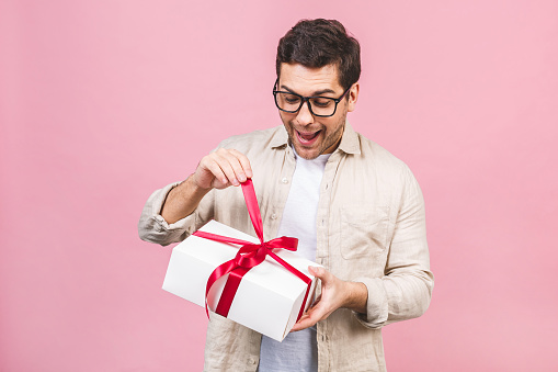 Holiday concept. Portrait of a young man opening gift box isolated over pink background.