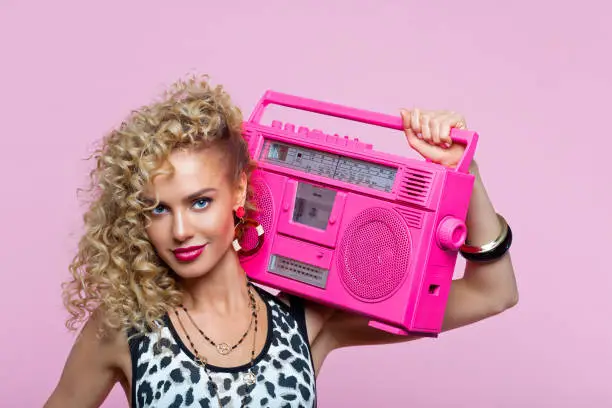 Summer portrait of beautiful long curly hair young woman wearing leopard print top and gold jewelry. Happy female in 80’s style outfit holding pink boom box on her shoulder, smiling at camera. Studio shot on pink background.