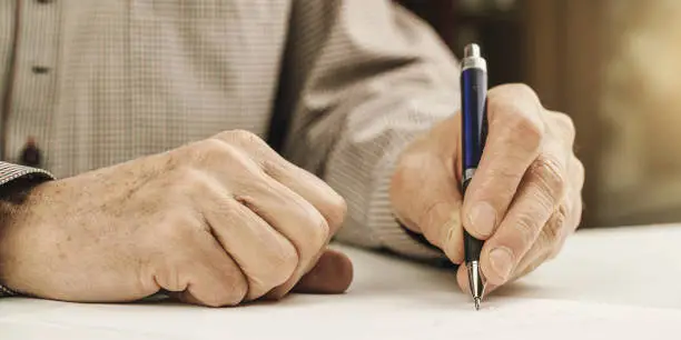 sick senior man in grey shirt tries to write letter with left hand using blue ball pen at table close view