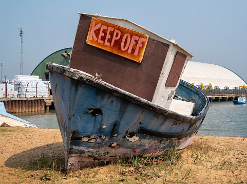 A ramshackle old boat stranded on the sandy beach at Harwich in Essex, Eastern England, on a sunny day. It bears a sign ordering the reader to “KEEP OFF”.