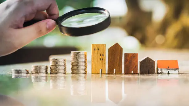 Photo of Hand holding magnifying glass and looking at house model with row of coin money, house selection, real estate concept.