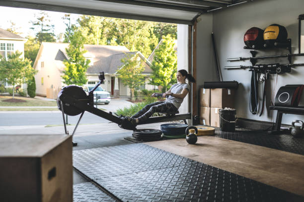 Active woman exercising on a rowing machine in her home garage gym during covid-19 pandemic. Photo of an active woman is exercising on a rowing machine in the home garage gym during covid-19 pandemic. Woman performing powerful pulls and focusing on endurance and cardio improvement. exercise room photos stock pictures, royalty-free photos & images