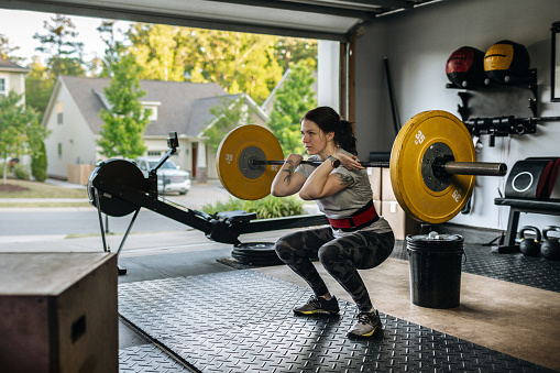 Photo of a fit woman performing a front squat with a heavy barbell in her home garage gym during covid-19 pandemic. Woman lifting a barbel and performing heavy powerlifting exercises. Power clean exercise. Focusing on core strength improvement.