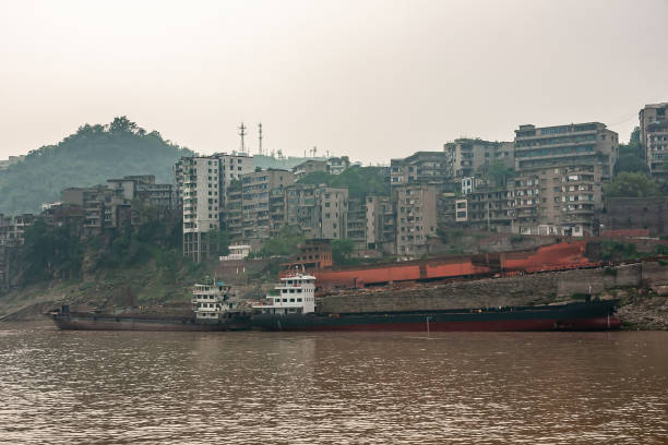 Scenic view of the Mekong River photographed from the old bridge, featuring docked boats along the riverbank, a colorful arched bridge in the distance, and modern city buildings surrounded by lush green hills, representing the harmony of nature, transportation, and urban development.