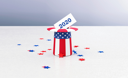 Patriotic American hat textured with American flag surrounded by star shaped confetti sitting on wood surface in front of defocused  background.  2020 written envelope entering the hat. Horizontal composition with copy space. Front view. 2020 Presidential Election concept.