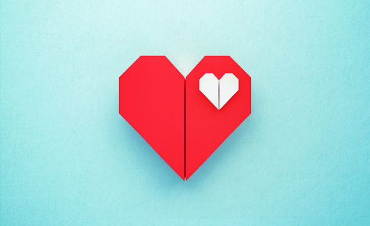 White origami heart sitting inside of a red origami heart on turquoise background. Horizontal composition with copy space. Love concept.