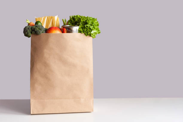 Paper bag with food gray background Various grocery items in paper bag on white table opposite gray wall. Bag of food with fresh vegetables, fruits, pasta and canned goods. Food delivery, shopping or donation concept. Copy space. groceries stock pictures, royalty-free photos & images