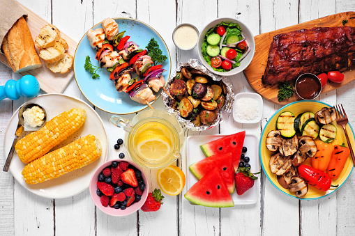 Summer BBQ or picnic food concept. Assortment of grilled meats, vegetables, fruits, salad and potatoes. Top down view table scene with a white wood background.