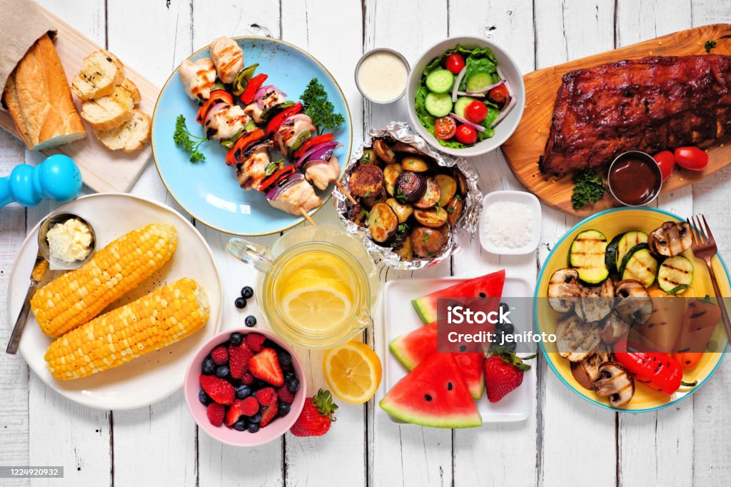 Summer BBQ or picnic food, top down view table scene over white wood Summer BBQ or picnic food concept. Assortment of grilled meats, vegetables, fruits, salad and potatoes. Top down view table scene with a white wood background. Summer Stock Photo