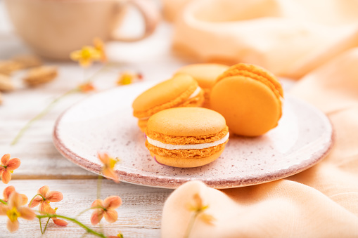 Orange macarons or macaroons cakes with cup of apricot juice on a white wooden background and orange linen textile. Side view, close up, selective focus.