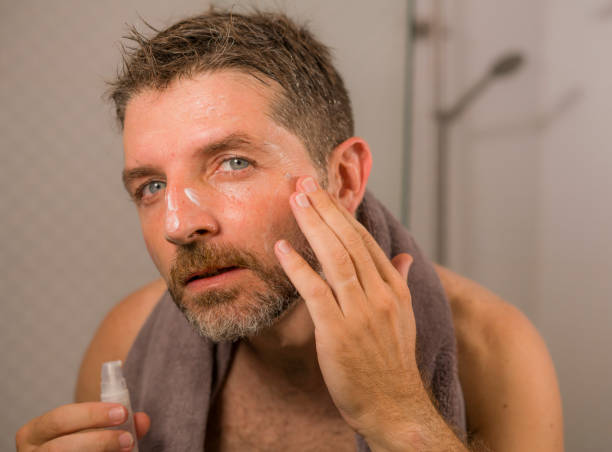 retrato de estilo de vida casero de joven hombre feliz y atractivo aplicando crema hidratante tratamiento de máscara facial mirando el espejo del baño en el cuidado de la piel anti envejecimiento masculino y concepto de belleza - male towel men human face fotografías e imágenes de stock