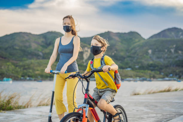 active school kid boy and his mom in medical mask riding a bike with backpack on sunny day. happy child biking on way to school. you need to go to school in a mask because of the coronavirus epidemic - child bicycle cycling danger imagens e fotografias de stock