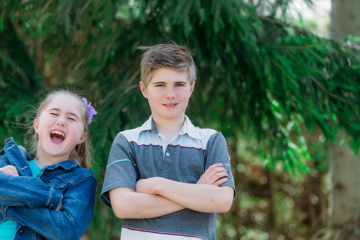 A serious confident boy has his arms crossed being standing beside his sister who is laughing. They are standing in front of a big spruce. She is wearing a jeans jacket. She has long blond hair tied. They are outside during spring in Quebec, Canada.