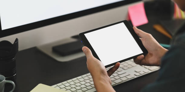 istock Cropped image of smart man hands holding a white blank screen computer tablet in front white blank screen computer monitor at the black working desk. 1224959173