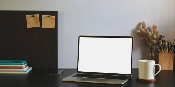 istock Photo of white blank screen computer laptop putting on black working desk and surrounded by stack of notebook, smartphone, pencil holder, wild grass in vase and coffee cup. 1224960120