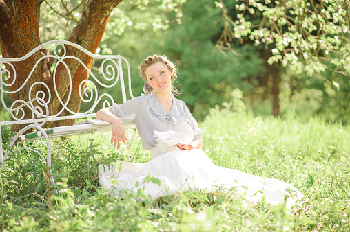 Young beautiful woman sits on the ground near a bench on a background of flowering apple trees. There is a book on the bench. She has a white dove in her hand.