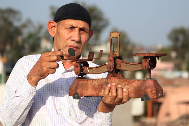 Old man of Indian Ethnicity carrying weight scale in hand outdoors. A balance is a device to measure weight or mass. These are also known as mass scales, weight scales, mass balances, weight balances, or simply scales, balances, or balance scales.