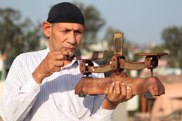 Old man of Indian Ethnicity carrying weight scale in hand outdoors. A balance is a device to measure weight or mass. These are also known as mass scales, weight scales, mass balances, weight balances, or simply scales, balances, or balance scales.