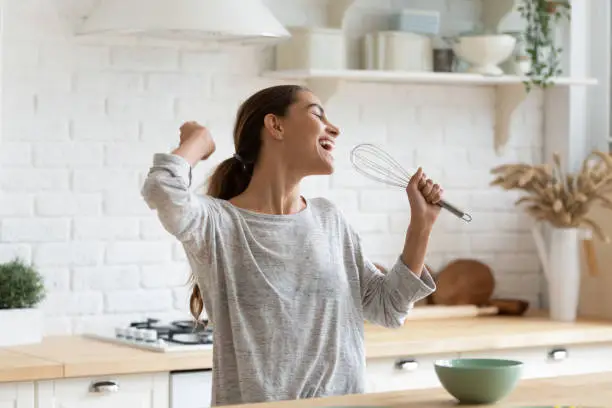 Photo of Excited funny girl singing into whisk, having fun in kitchen