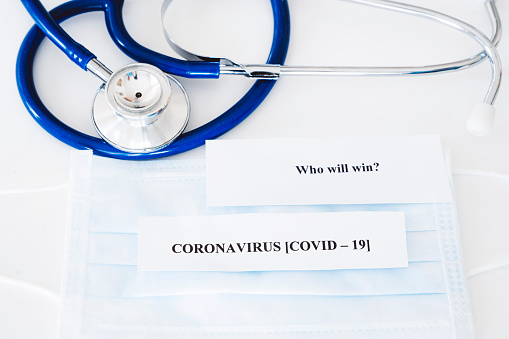 Close-up photo of face mask and stethoscope on a white background. Note on the mask saying Who will win? - Corona virus (covid-19) . Global pandemic of Covid-19 (Coronavirus).