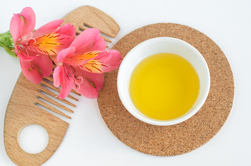 Small white bowl with cosmetic oil, red flowers and wooden hairbrush. Natural spa and beauty treatment recipe. Top view, copy space.